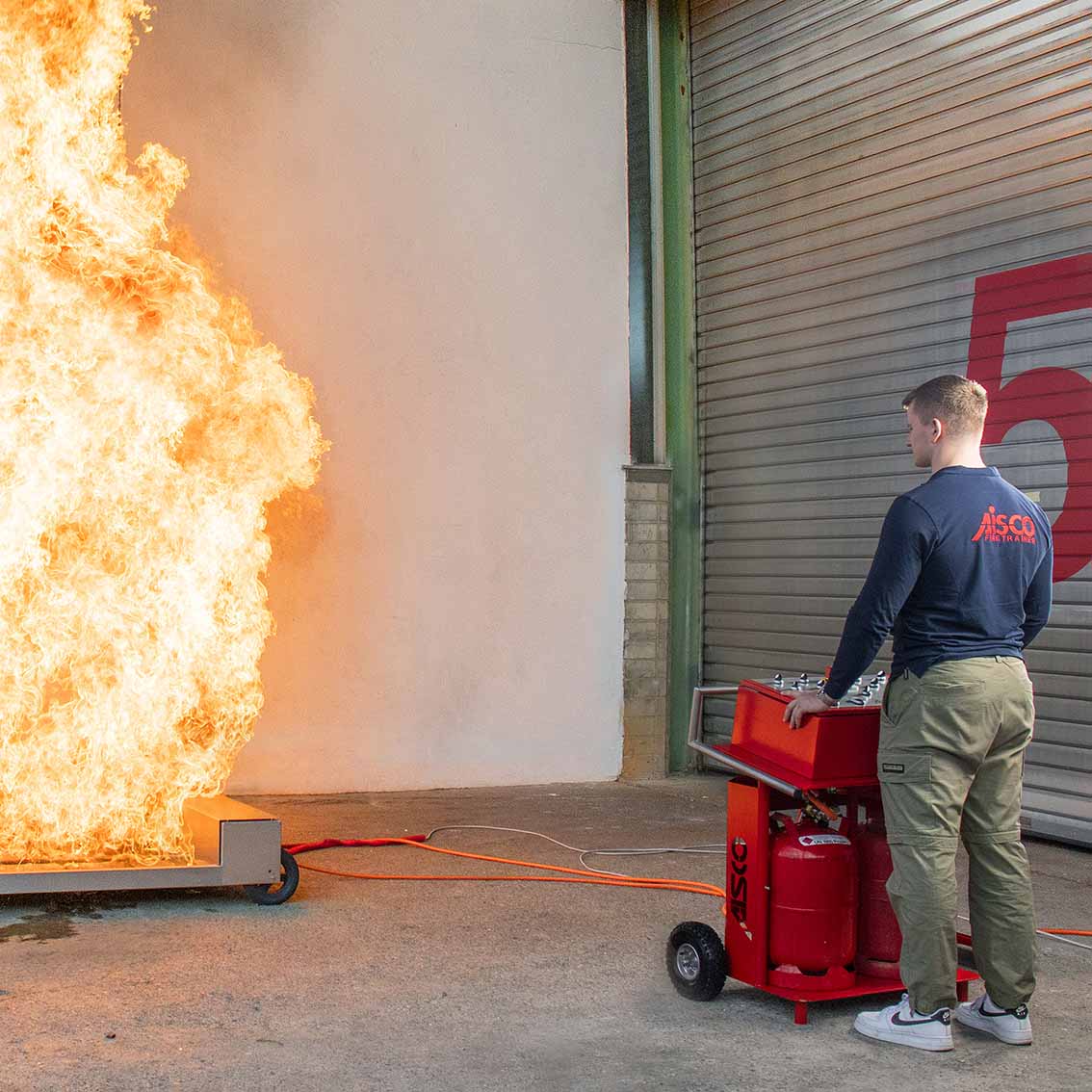 Eine Person steuert über das AISCO Bedienpult F 4 den Firetrainer E1502 XL, der mit meterhohen Flammen brennt – realitätsnahe Feuerwehrübung mit 4-Kanal-Steuerung