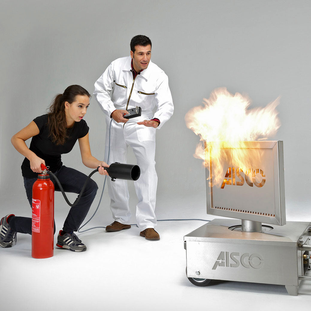 Eine Person löscht einen Elektrobrand am AISCO Firetrainer Caddy M, während der Brandschutzunterweiser daneben Anweisungen gibt. Der brennende Caddy M ist mit Monitor-Aufsatz sichtbar.