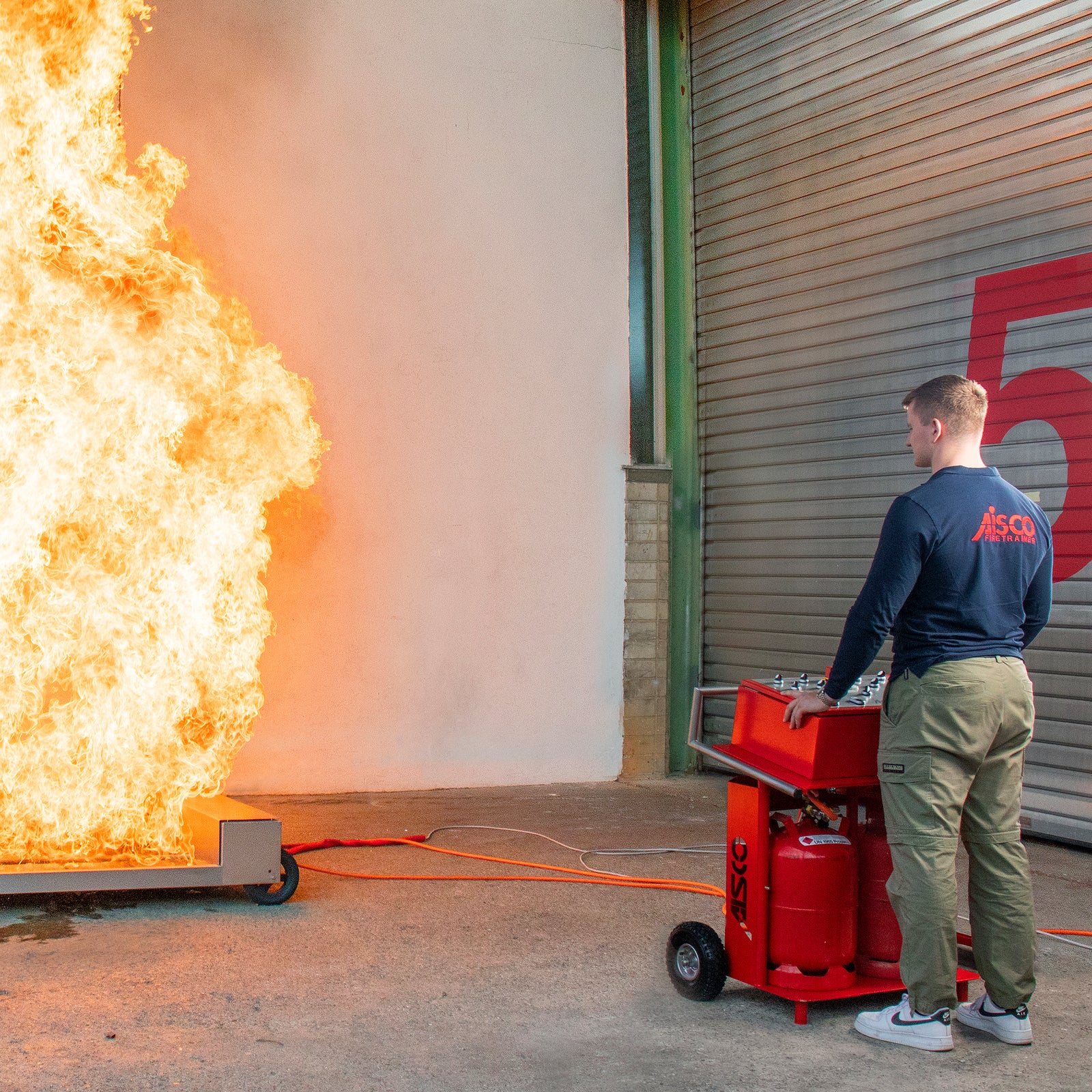 A person controls the Firetrainer E1502 XL, which is burning with meter-high flames, via the AISCO control panel F 4 – a realistic fire department exercise with 4-channel control